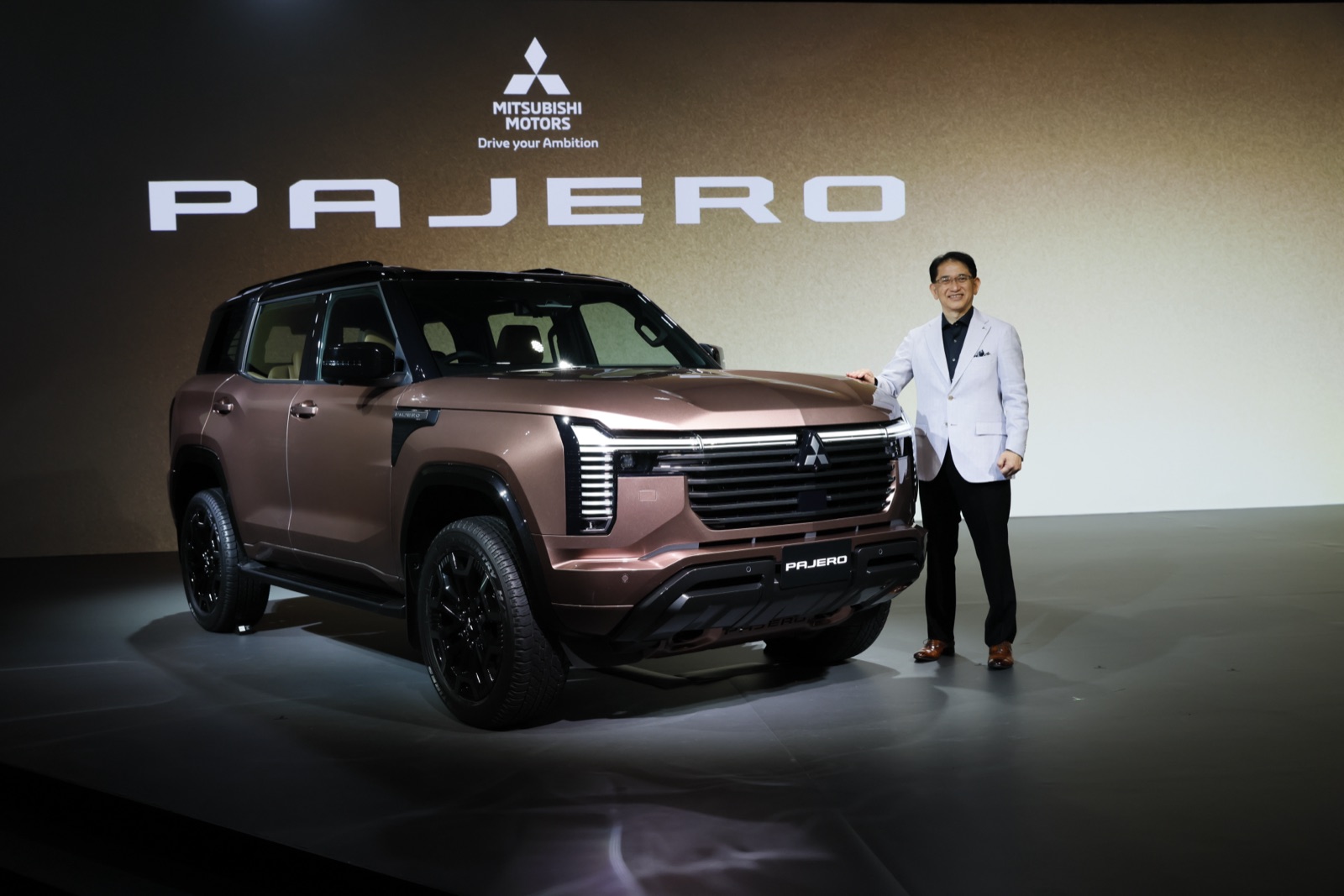 The new Mitsubishi Pajero on stage at its reveal, with the PAJERO name illuminated on the wall behind the bronze SUV.